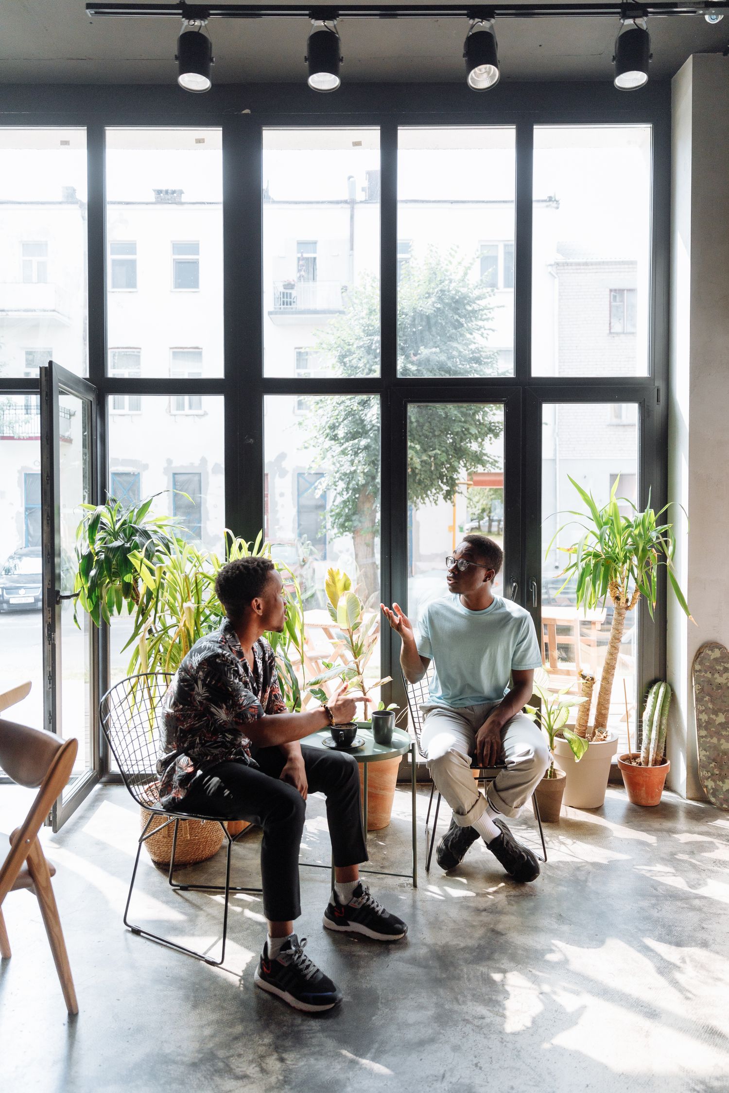Two men engaged in conversation in a bright, plant-filled urban cafe.