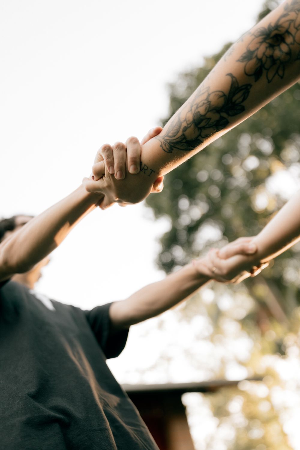 Two tattooed arms holding hands outdoors, symbolizing connection and unity.